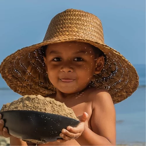 Smiling child with a sun hat holding a bowl of sand on the beach at a family-friendly resort in Lombok