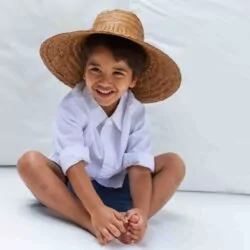 Smiling child in a white shirt and straw hat sitting playfully at The Lombok Lodge, a luxury family resort in Lombok